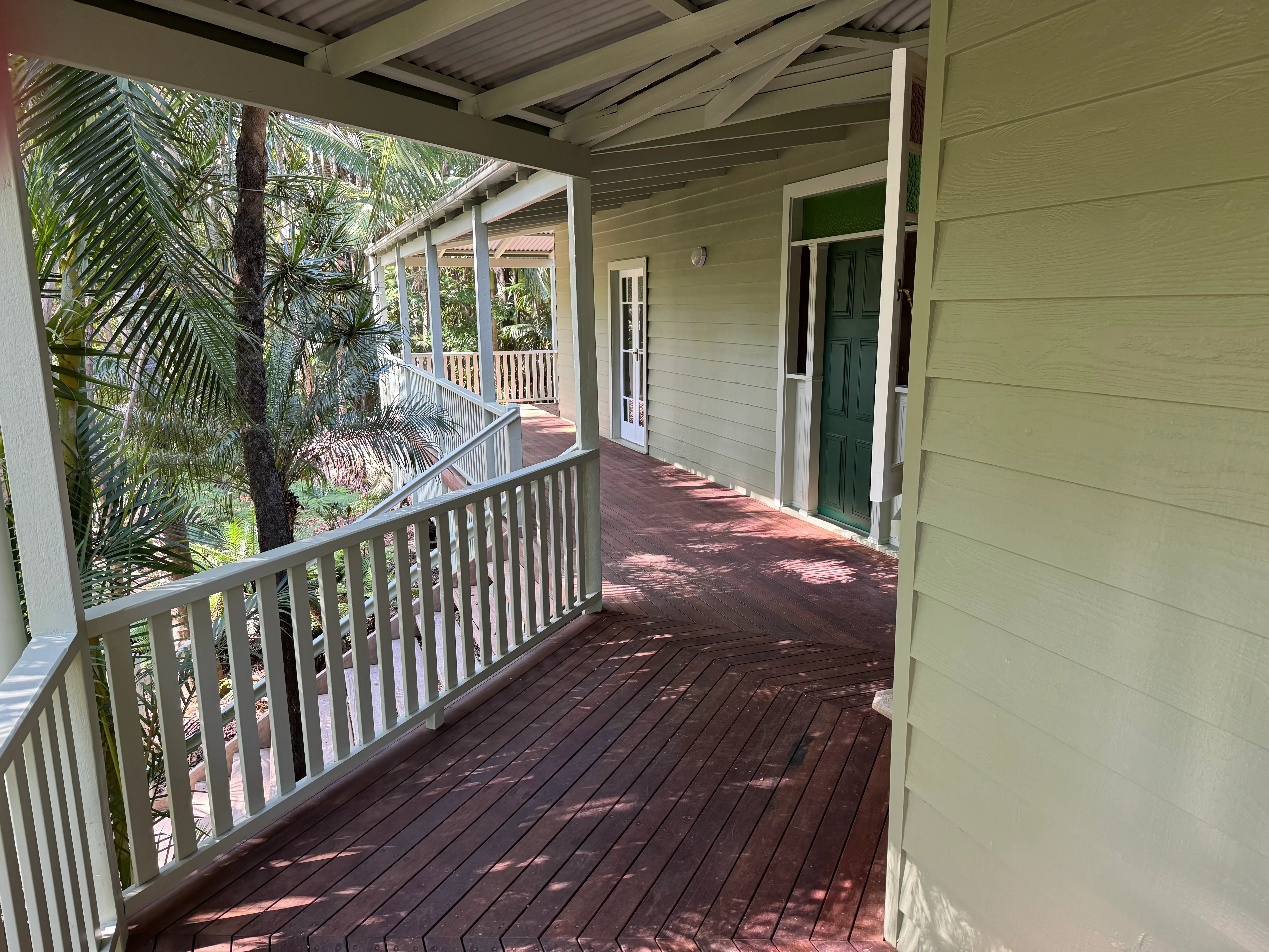 Painted timber verandah of a weatherboard home, with balustrade, timber decking and a dark green front door, surrounded by greenery.