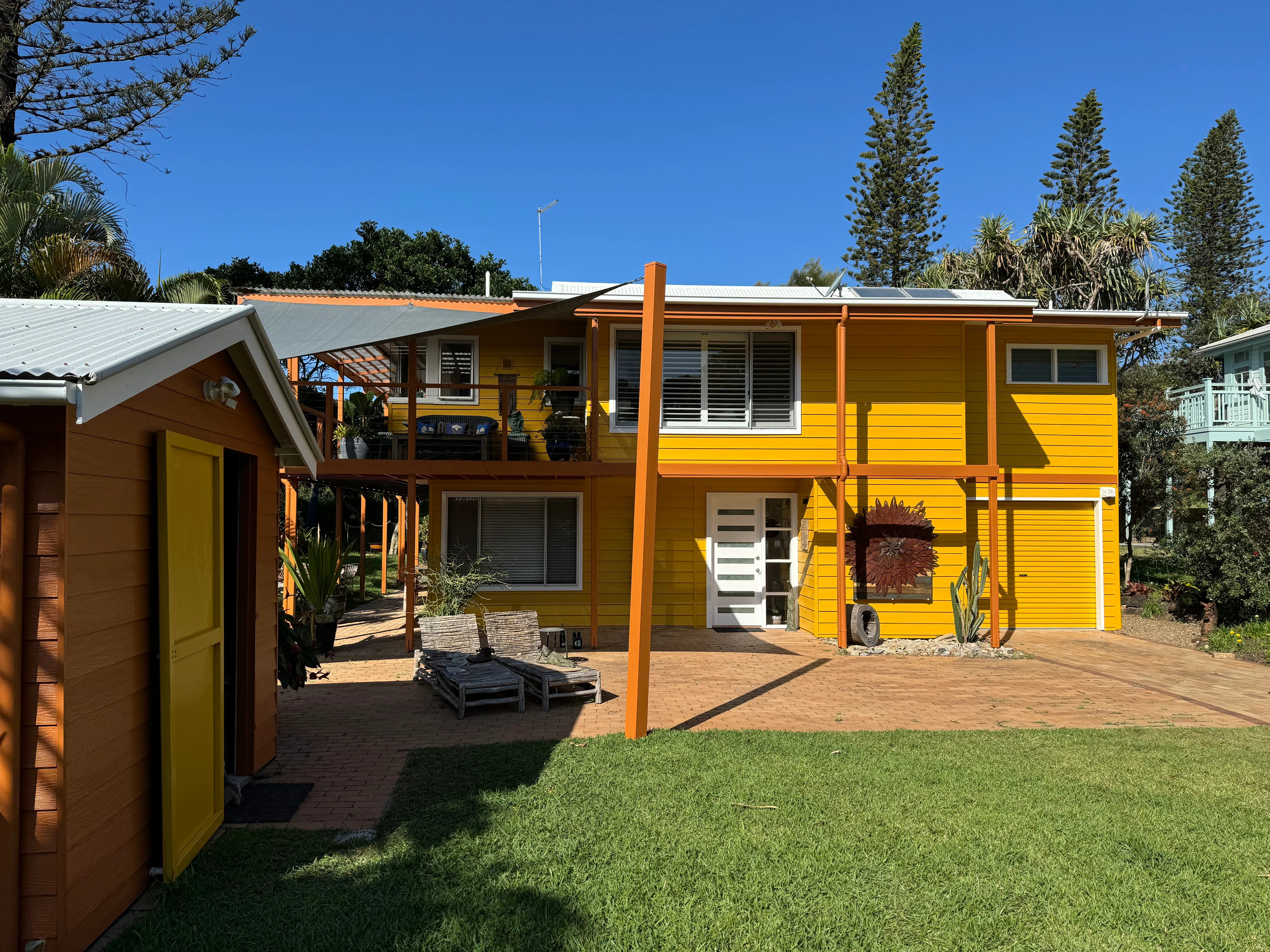 Two-storey weatherboard home painted yellow with orange trim, a white entry door and garage door, beside a matching timber shed.